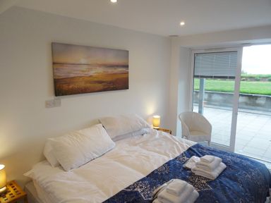 A bedroom with a bed covered with white linens and blue blanket with folded towels a painting on the wall and a glass door leading outside at The Sandcastle in Freshwater East