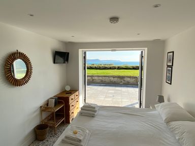 A bedroom with a bed towels and a TV facing a grass lawn and sea visible through open doors at The Sandcastle in Freshwater East