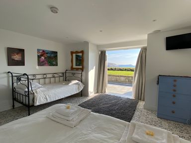 A bedroom with a bed a daybed a blue dresser and a sliding door with curtains showing a lawn and hills at The Sandcastle in Freshwater East