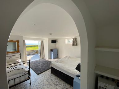A bedroom with two beds a blue dresser a mirror a rug a tv and an open door showing grass and sea at The Sandcastle in Freshwater East