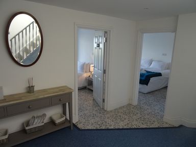 A hallway with a console table and an oval mirror showing stairs and two doorways leading to bedrooms at The Sandcastle in Freshwater East
