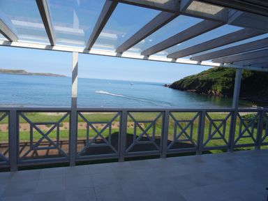 A balcony with a railing and glass roof overlooking the sea and a grassy shore at The Sandcastle in Freshwater East