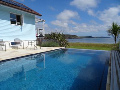 A swimming pool next to a blue house with chairs and a table overlooking the sea at The Sandcastle in Freshwater East