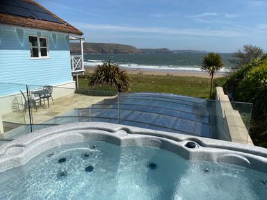 A hot tub on a patio overlooking a lawn and a beach with a blue house and outdoor table and chairs at The Sandcastle in Freshwater East