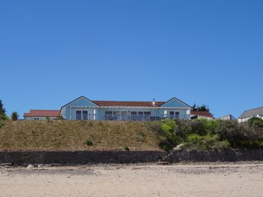 A house on a raised area with grass and bushes in front of a sandy beach at The Sandcastle in Freshwater East