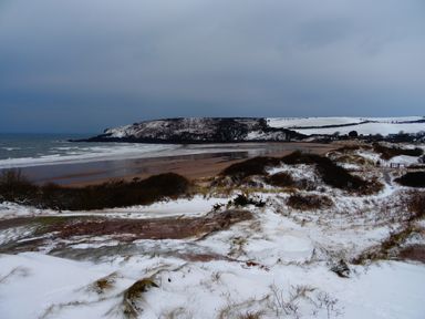 A snow-covered beach with waves hitting the shore and a headland in the distance at The Sandcastle in Freshwater East