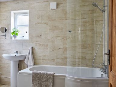 A bathroom with a bathtub shower combination a pedestal sink a small window with a plant a towel hanging and beige tiled walls at Lilly Cottage in Whitecroft Royal Forest of Dean