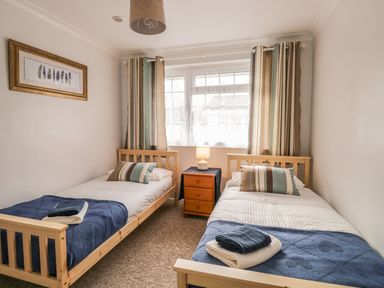 A bedroom with two single beds, striped pillows, blue blankets, a wooden nightstand with a lamp and striped curtains at Ducks Ditty in Liskeard