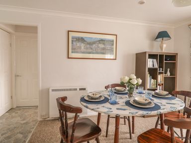 A dining room with a table set for four with a floral tablecloth and wooden chairs and a wooden shelving unit with books and a lamp at Ducks Ditty in Liskeard