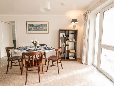 A dining area with a round table set with plates and glasses four wooden chairs a bookshelf with a lamp and a large window with curtains at Ducks Ditty in Liskeard