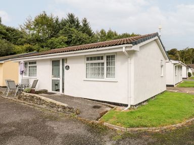 A white single-story house with outdoor table and chairs on a paved area surrounded by grass at Ducks Ditty in Liskeard