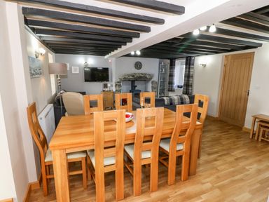 A dining area with a wooden table and eight chairs next to a living room with sofas a television and a stone fireplace at Min-y-rhos in Edern