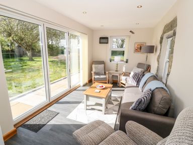 A sitting room with grey sofa and armchairs a wooden coffee table and large sliding glass doors showing a garden at Min-y-rhos in Edern