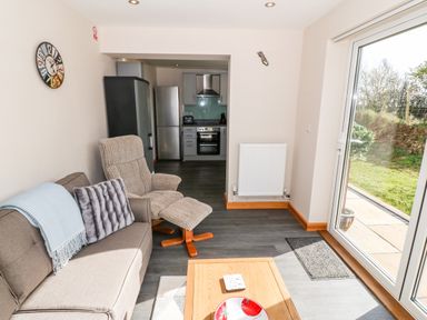 A living room with a sofa armchair and coffee table next to a kitchen and patio doors showing a garden at Min-y-rhos in Edern