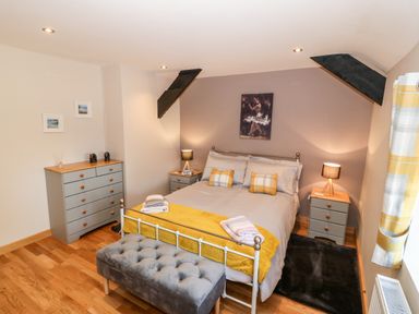 A bedroom with a bed with yellow bedding and pillows two bedside tables with lamps a grey dresser and a grey padded bench at Min-y-rhos in Edern