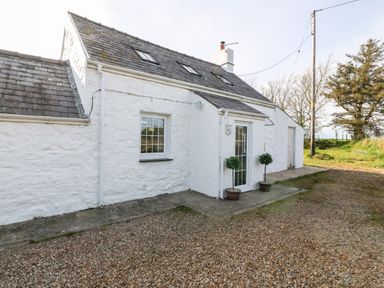 The exterior of a white stone cottage with a gravel driveway and two potted plants near a glass door at Min-y-rhos in Edern