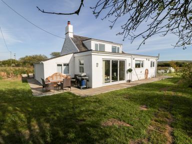 A white house with a chimney a patio with outdoor furniture and a barbecue grill in a grassy yard at Min-y-rhos in Edern
