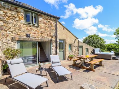 An outdoor stone patio with two lounge chairs a small table a wooden picnic table and a hot tub at Sunbury Barn in St Agnes