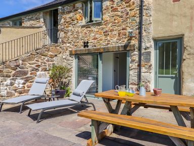 A stone exterior patio with two lounge chairs and a wooden picnic table at Sunbury Barn in St Agnes