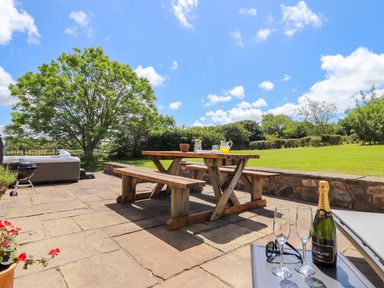 An outdoor patio with a wooden picnic table and benches a bottle of champagne with glasses a barbecue and a hot tub in a garden at Sunbury Barn in St Agnes