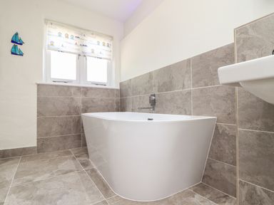 A bathroom with tiled walls and floor a white bathtub and a window with a patterned blind at Sunbury Barn in St Agnes