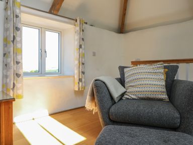 A sitting area with a grey armchair a patterned cushion a window with curtains and wooden floor at Sunbury Barn in St Agnes