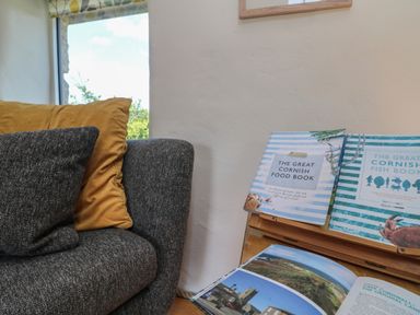 A corner of a room with a grey couch with cushions near a wooden stand holding cookbooks at Sunbury Barn in St Agnes