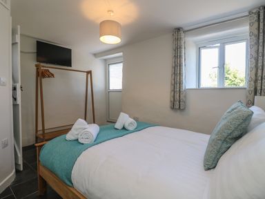 A bedroom with a bed covered with white linens and blue throw and pillows two windows with patterned curtains a wooden clothes rack with hangers and a wall-mounted TV at Sunbury Barn in St Agnes