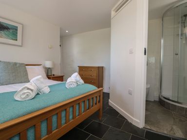 A bedroom with a wooden bed with rolled towels on a blue cover a wooden dresser a bedside table with a lamp and a bathroom with a glass shower at Sunbury Barn in St Agnes