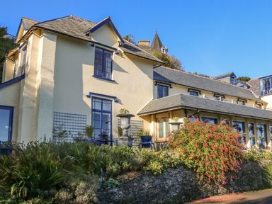 A building with beige walls and blue window frames surrounded by plants and flowers at C S Lewis Balcony in Lynton
