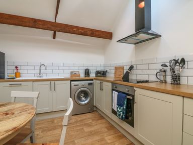 A kitchen with white cabinets wooden countertops a washing machine an oven a round wooden table and kitchen utensils at C S Lewis Balcony in Lynton