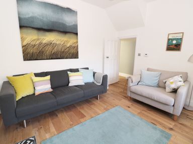 A living room with a dark gray sofa and light gray armchair with pillows a blue rug on the floor and artwork on the walls at Teviot Cottage in Corbridge