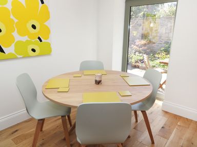 A dining area with a round wooden table and four light gray chairs near a glass door and yellow flower artwork on the wall at Teviot Cottage in Corbridge