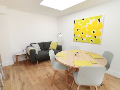 A dining area with a round wooden table set with yellow placemats and light grey chairs next to a dark grey sofa with cushions and a side table at Teviot Cottage in Corbridge