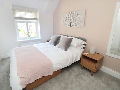 A bedroom with a bed with white bedding and gray pillows a wooden nightstand with a lamp a window with blinds and a painting on the wall at Teviot Cottage in Corbridge