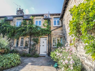 A stone cottage with white windows and door surrounded by green plants and flowers at Teviot Cottage in Corbridge