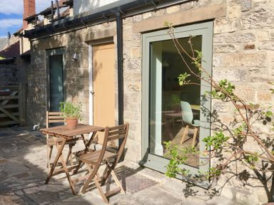 A stone exterior wall with a wooden door and large window with frame and a small wooden table and two chairs outside with a potted plant on the table at Teviot Cottage in Corbridge