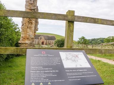 A wooden fence and an information board about Abbotsbury Abbey with ruins and countryside in the background at Abbotsbury Abbey in Abbotsbury