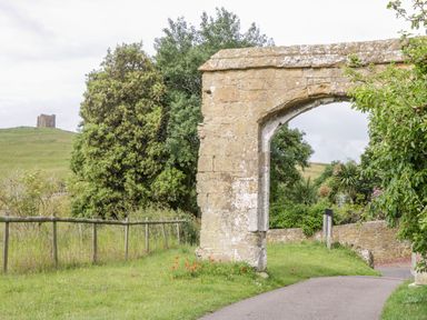 A stone archway next to a paved path with trees and a hill with a small stone structure in the background at Market House in Abbotsbury