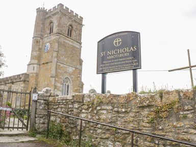 A stone church tower with a clock and a sign for St Nicholas Abbotsbury behind a stone wall at Market House in Abbotsbury