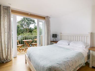 A bedroom with a double bed a lamp on a bedside table and balcony doors opening to outdoor wooden chairs and table at Crift Farm Cottage Lanlivery near Lostwithiel