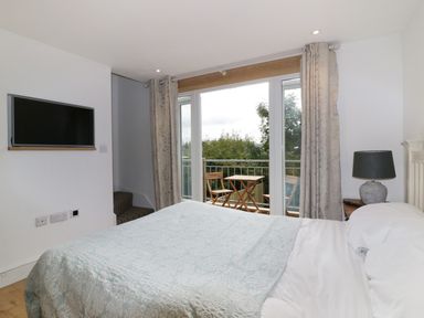 A bedroom with a bed a bedside table with lamp and a TV on the wall with glass doors opening to a balcony with a table and chairs at Crift Farm Cottage Lanlivery near Lostwithiel