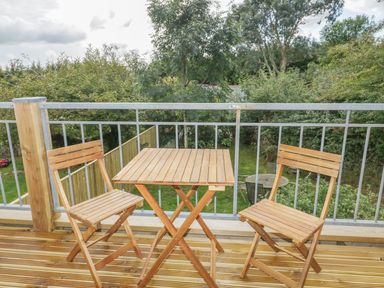 A wooden table with two wooden chairs on a balcony overlooking a green garden at Crift Farm Cottage in Lanlivery near Lostwithiel