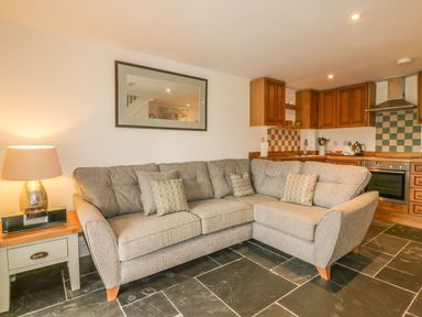 A living room with a gray sectional sofa and a lamp on a side table in an open kitchen with wooden cabinets and tiled backsplash at Crift Farm Cottage Lanlivery near Lostwithiel