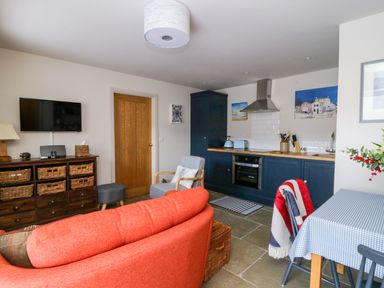 A living area with an orange sofa a grey armchair a wooden cabinet with baskets a wall-mounted TV and a kitchen with blue cabinets at Wallerthwaite Barn Cottage in Markington