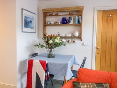 A dining area with a checkered tablecloth on the table a vase with flowers on the table a wall shelf with mugs teapots and books and a wooden door at Wallerthwaite Barn Cottage in Markington