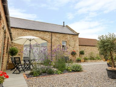 An outdoor patio with a table umbrella chairs and purple flowers in front of a stone building at Wallerthwaite Barn Cottage in Markington