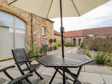 An outdoor patio with a black wooden table and chairs under a large umbrella next to a stone building with flowers at Wallerthwaite Barn Cottage in Markington