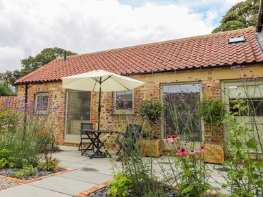 A patio area with a table umbrella and chairs outside a brick cottage with flowers in the garden at Wallerthwaite Barn Cottage in Markington