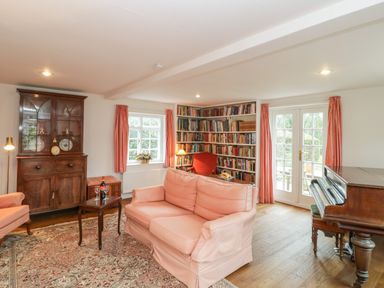 A living room with a pink sofa armchairs a wooden cabinet bookshelf a piano and French doors at Church View in Salisbury